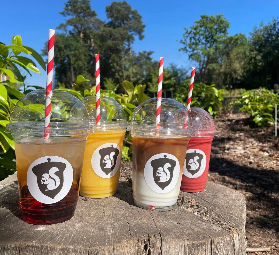 Four iced drinks in clear plastic cups with red and white straws on a wooden surface outdoors.