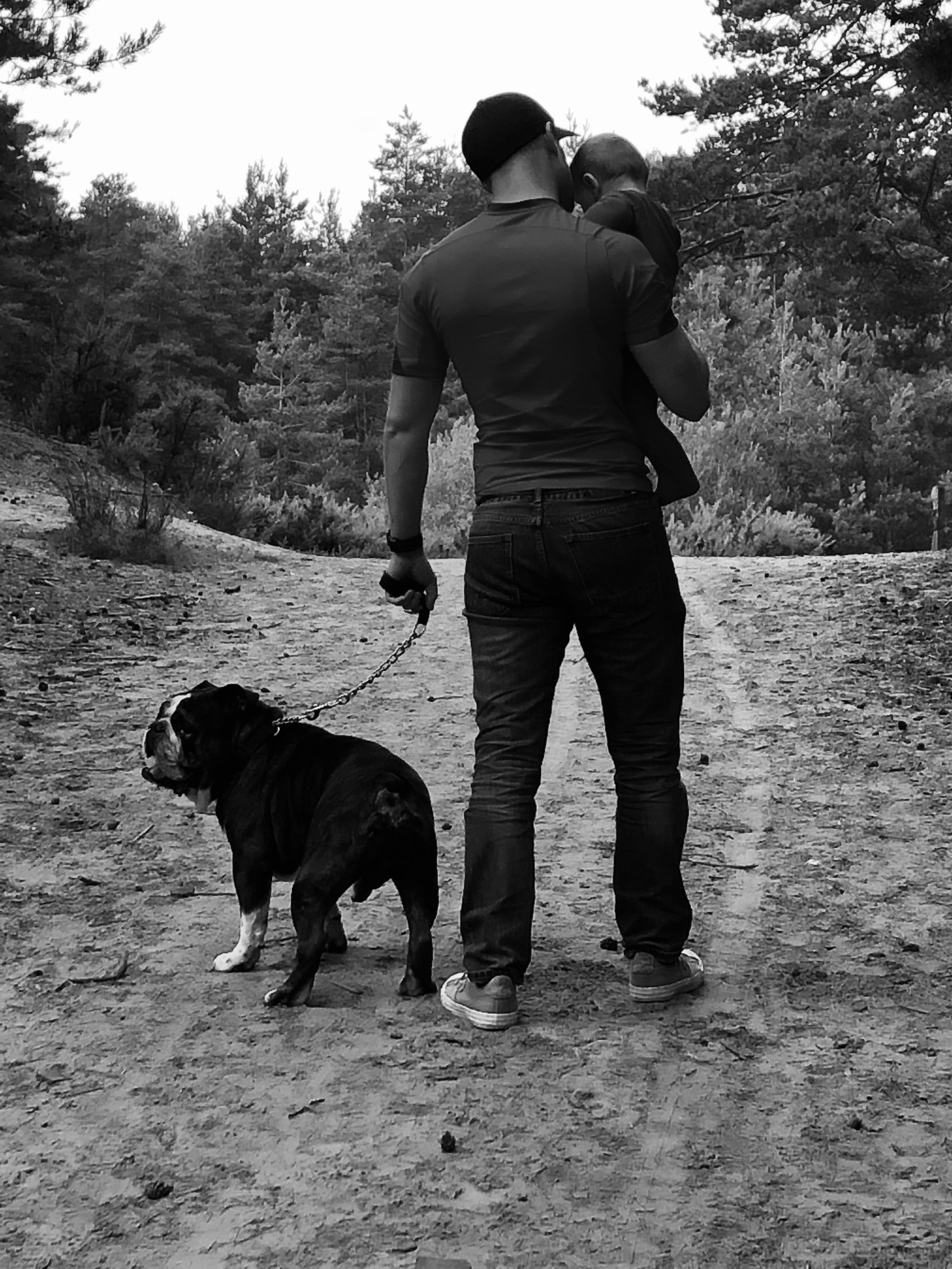 A black and white lifestyle photograph of a man standing on a dirt path in a wooded area, holding a child on his shoulders and wearing a chain leash next to a large black dog.